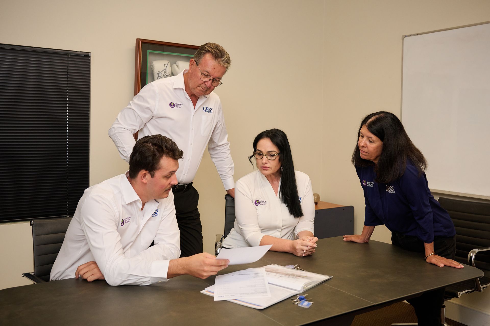Four people in a meeting around a table. One man points to papers. Others listen intently, one leans in.