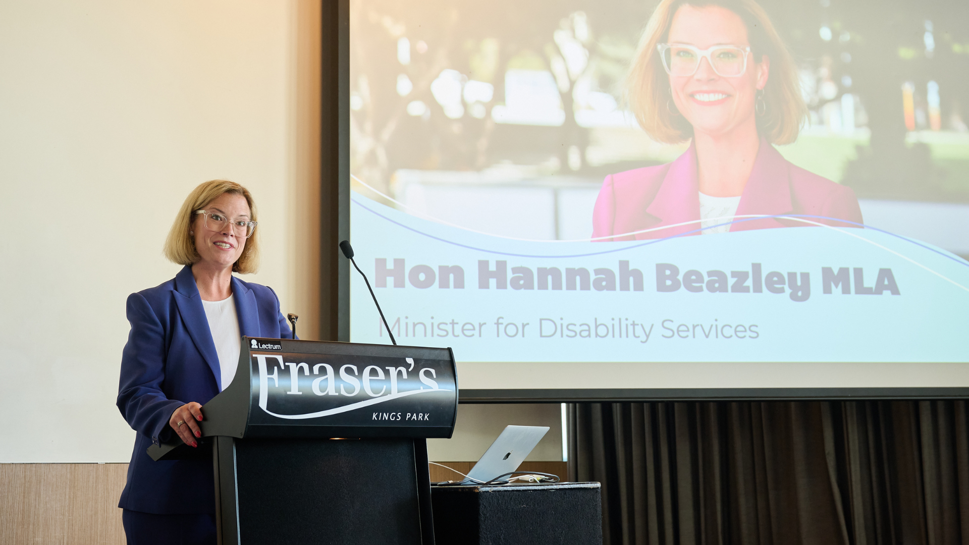 Woman at a podium, speaking at Frasers, with her photo and title on a screen behind. She wears a blue suit.