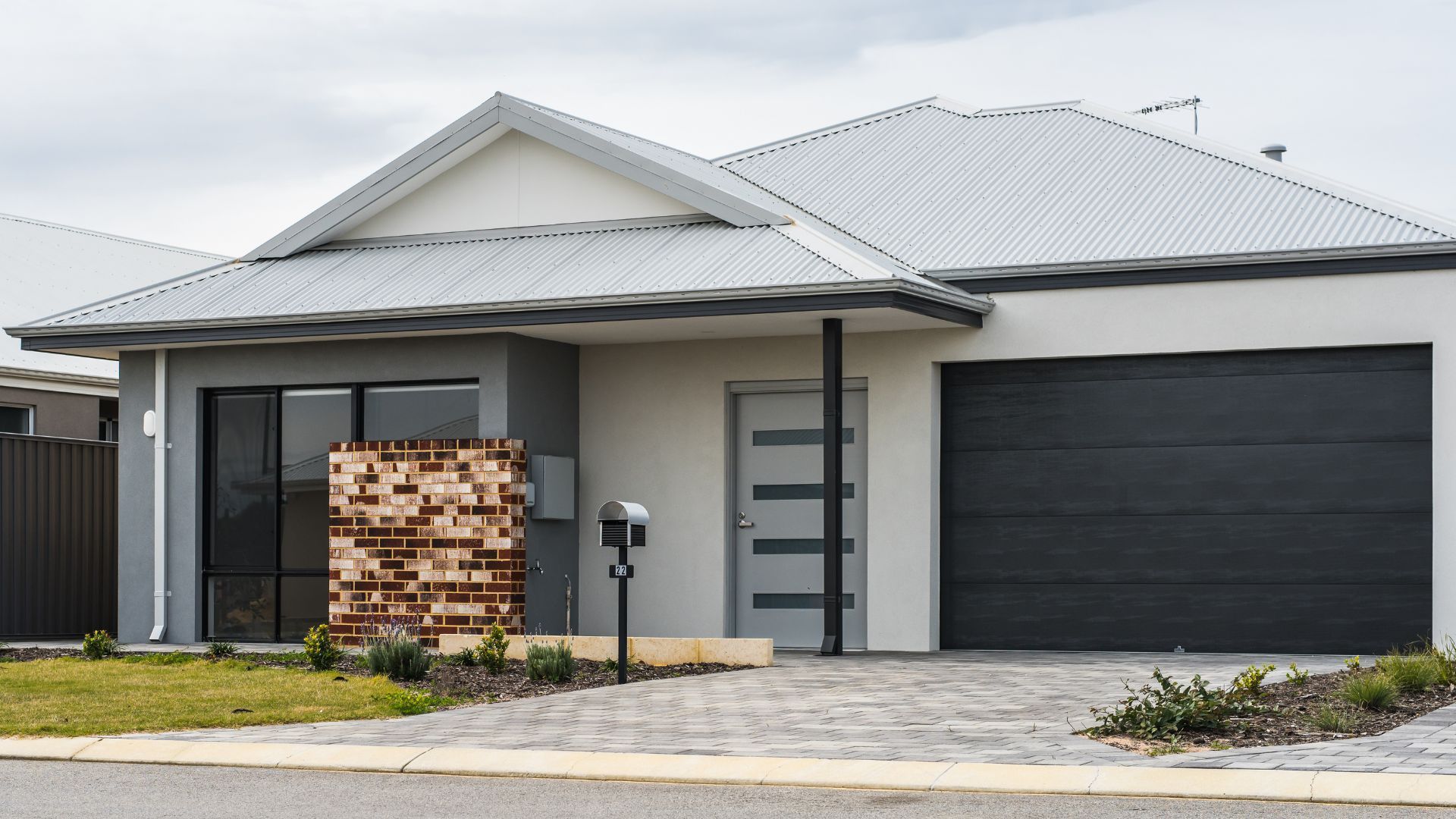 Modern single-story house with gray siding, garage, and patterned brick wall. Overcast sky.