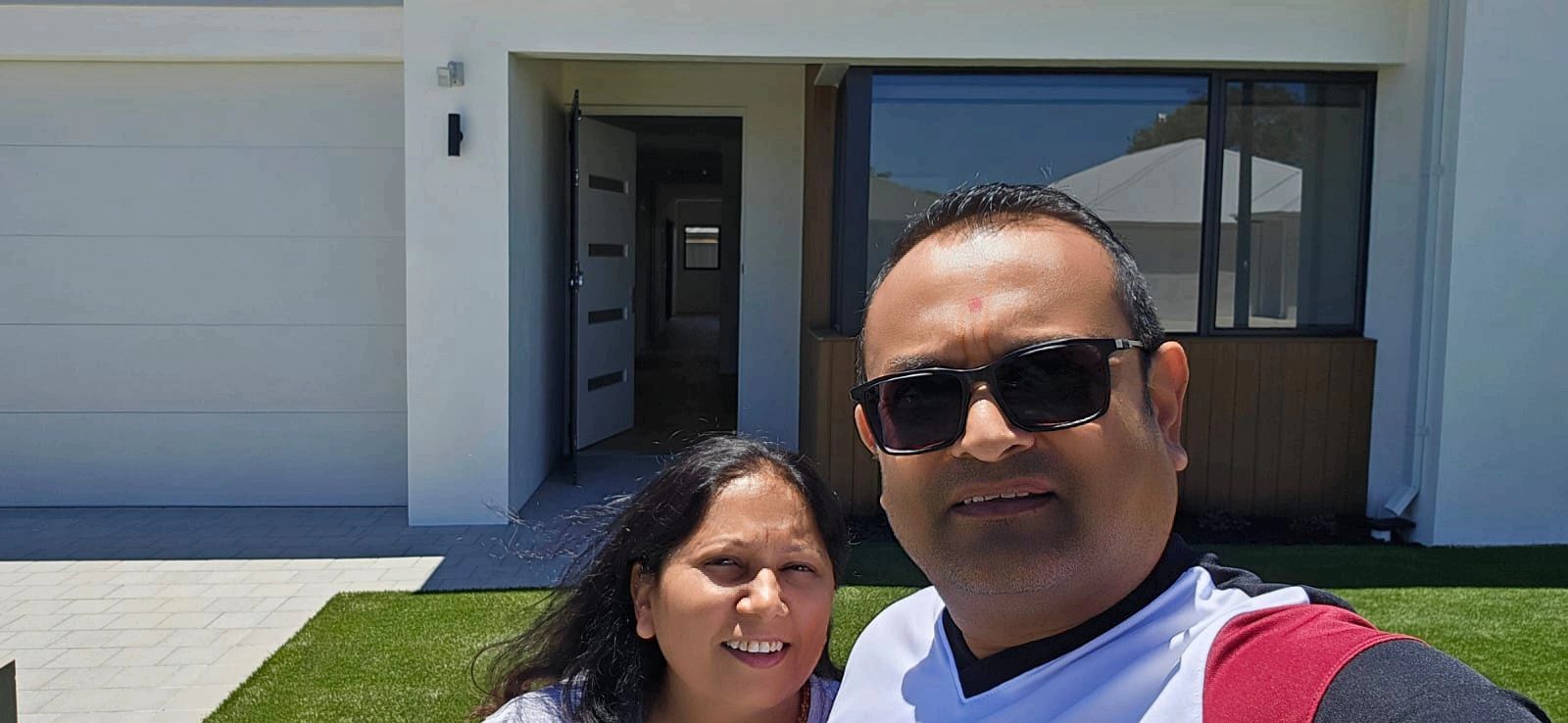 A person wearing sunglasses and another person stand smiling in front of a modern white house with a garage and open door.