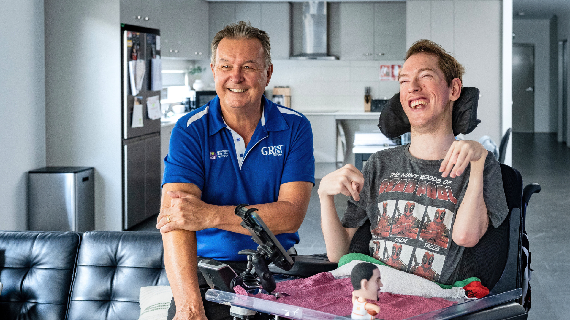 Man and young person in wheelchair smiling.  Man's arm is resting on wheelchair. Home interior, with kitchen in background.