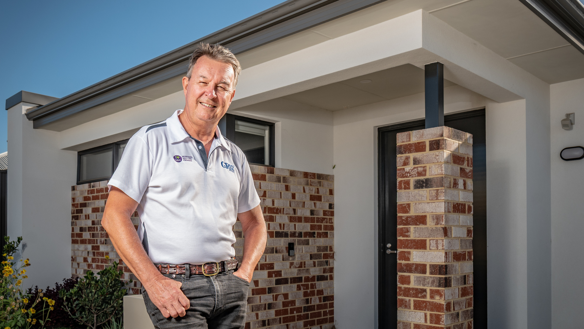 Man in front of a house, smiling with hands in pockets. White top, patterned pants. Brick and white exterior.