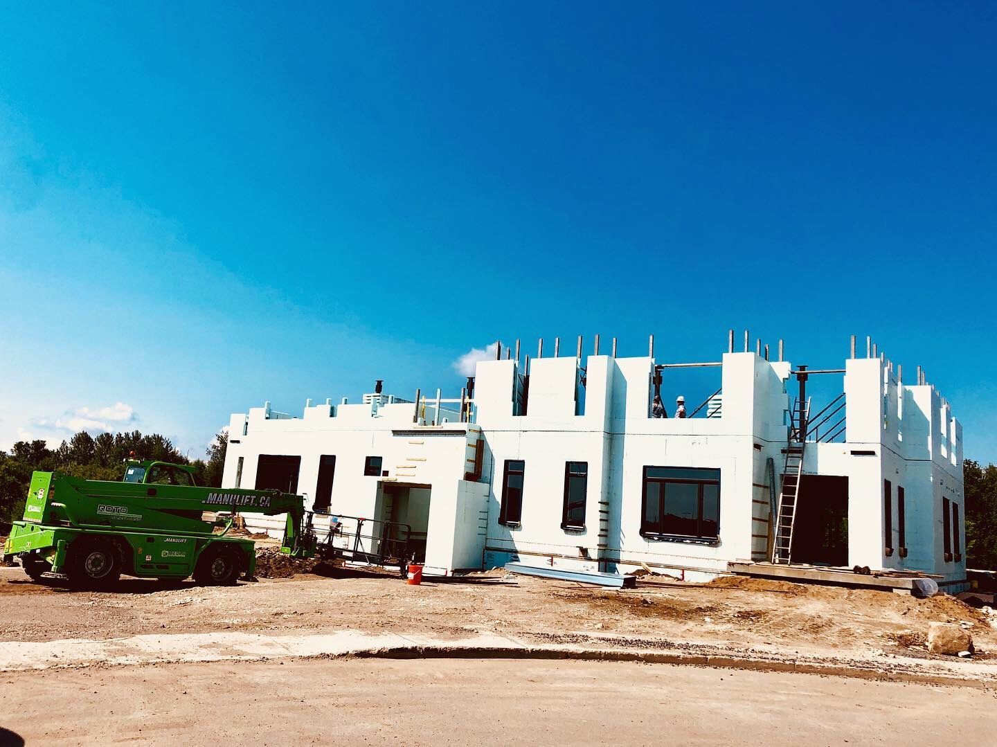 Chantier d'un bâtiment blanc aux fenêtres ouvertes sous un ciel d'un bleu éclatant. Un chariot élévateur vert est visible.