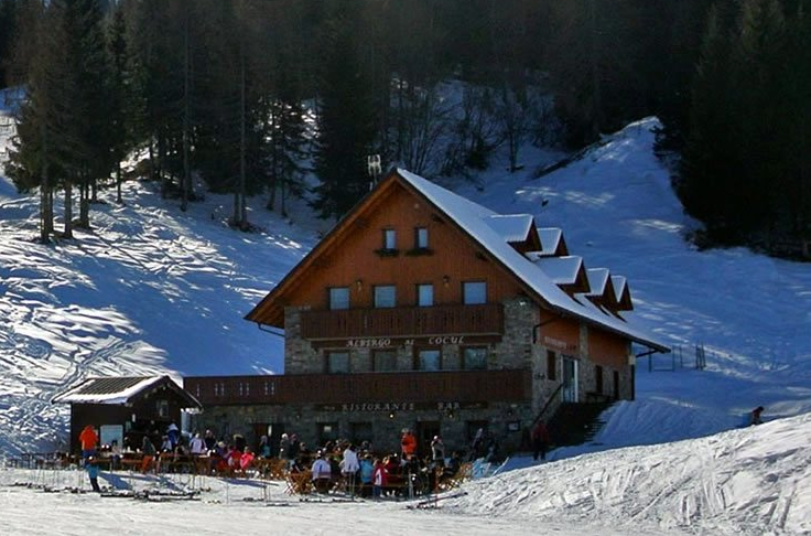 Rifugio di montagna innevato con persone all'esterno; alberi e piste da sci sullo sfondo. Giornata di sole.