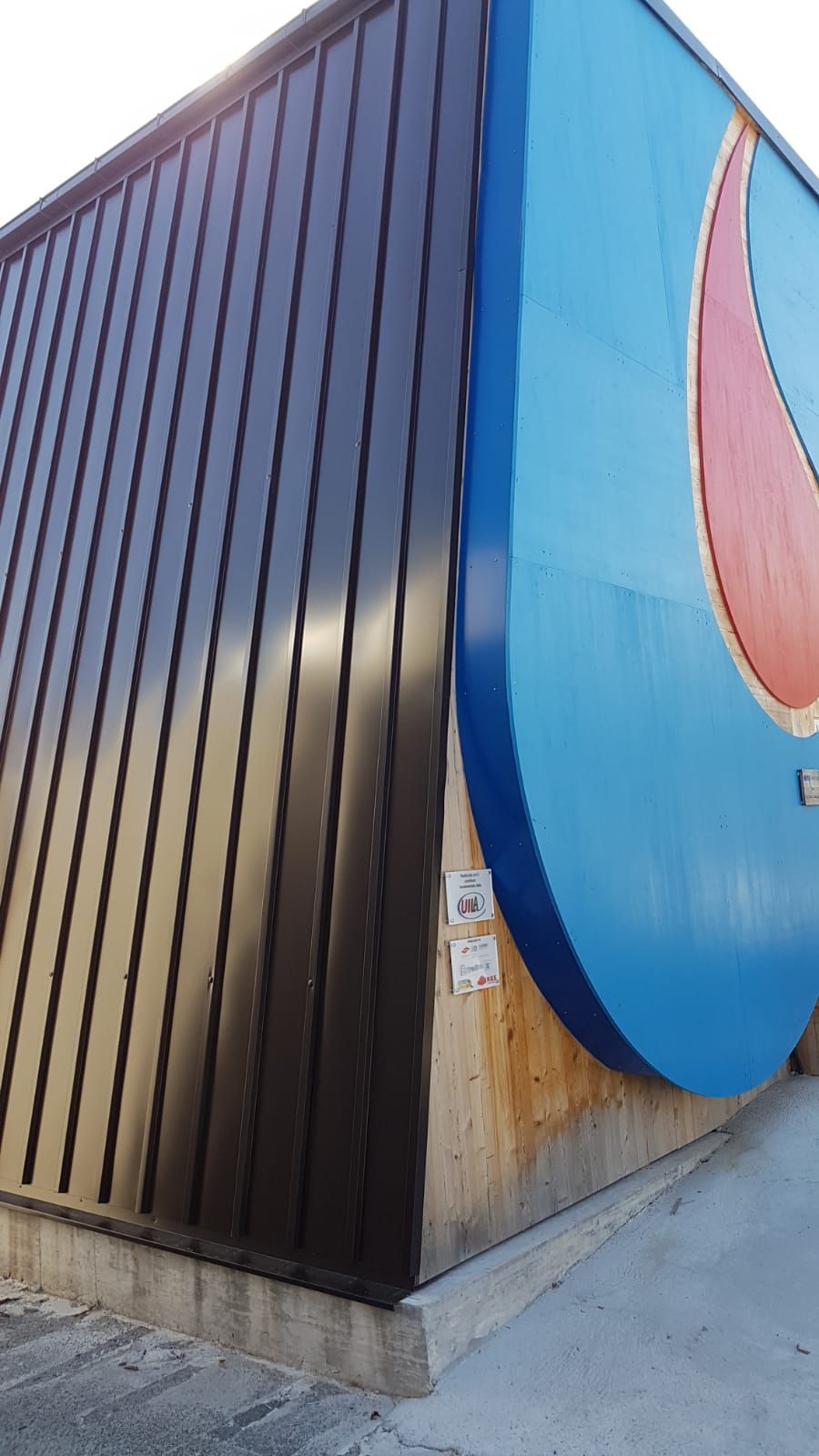 Angolo di un edificio con rivestimento in metallo nero e logo Pepsi blu, base in cemento.