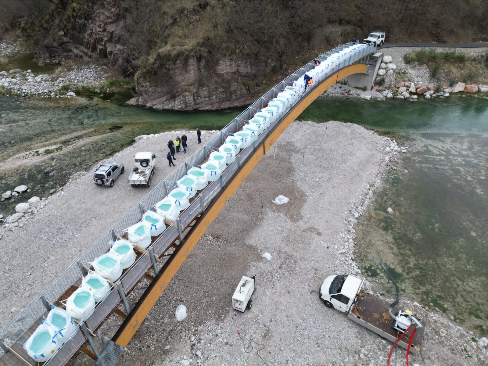 Borse allineate su un ponte, persone e veicoli ai lati. Vista su un fiume tra le rocce.