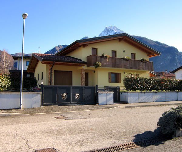 Casa gialla con finiture e balcone marroni, cancello nero, montagne sullo sfondo.