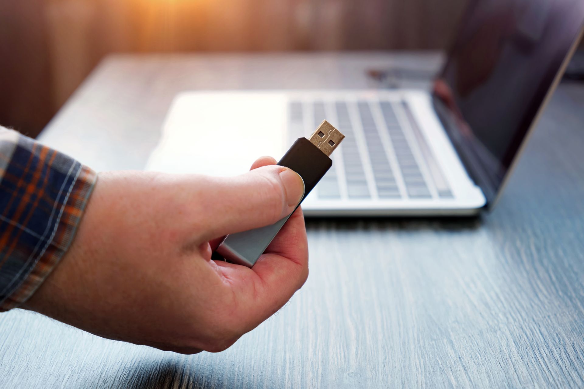 Hand holding a USB flash drive near an open laptop on a wooden desk.