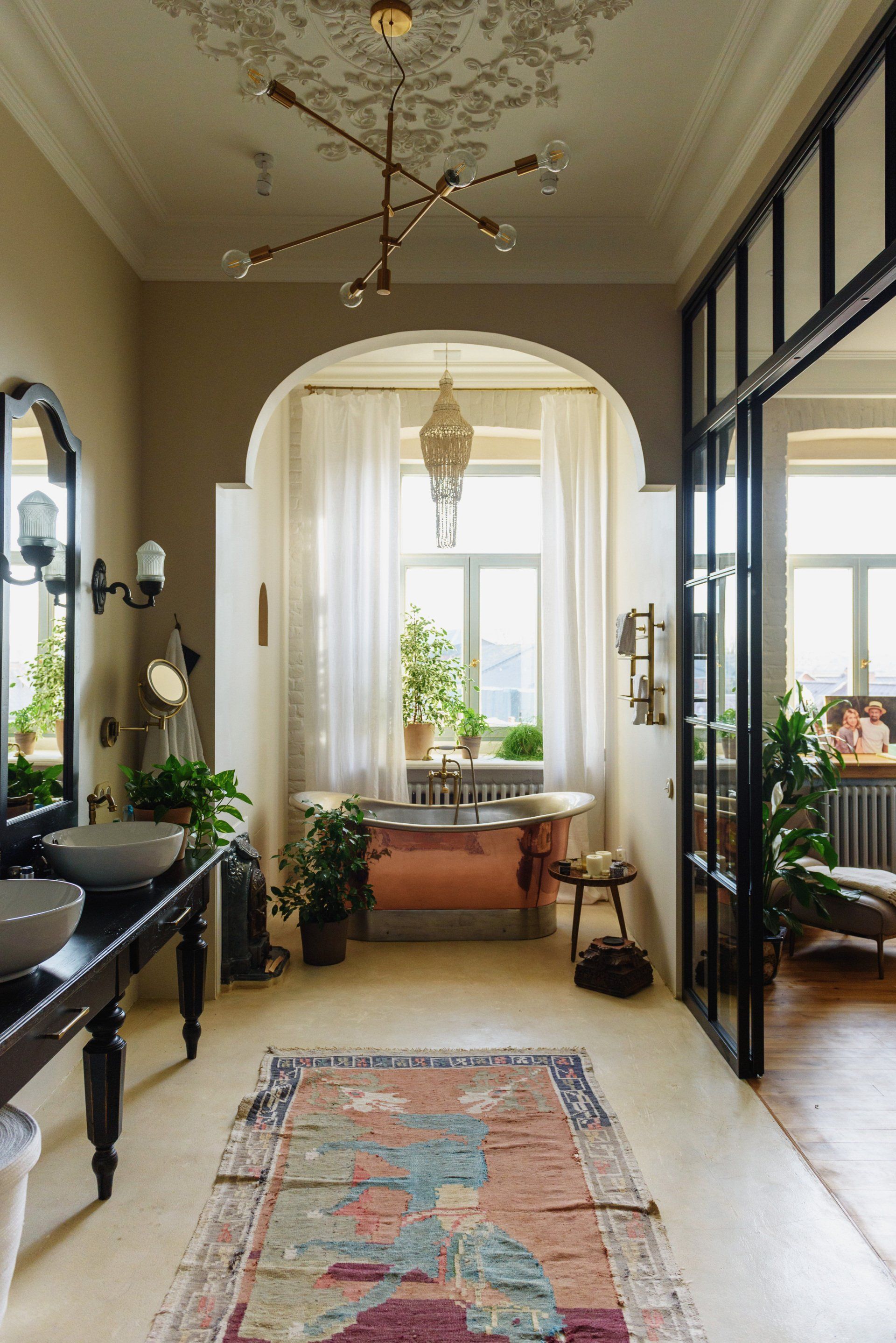 A spacious, ornate bathroom features a copper bathtub under an arched window, a black vanity, and a vintage-style rug.