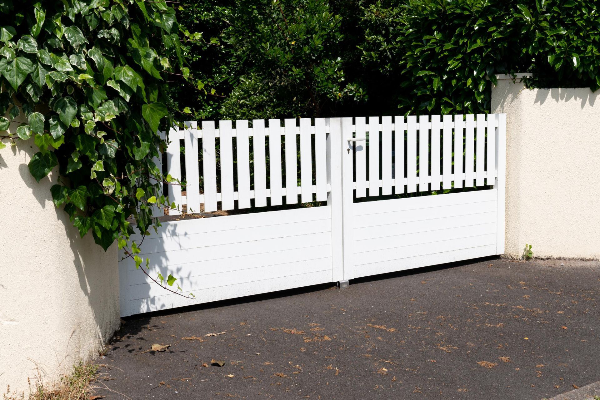 White picket gate in a driveway, surrounded by a wall and green foliage.