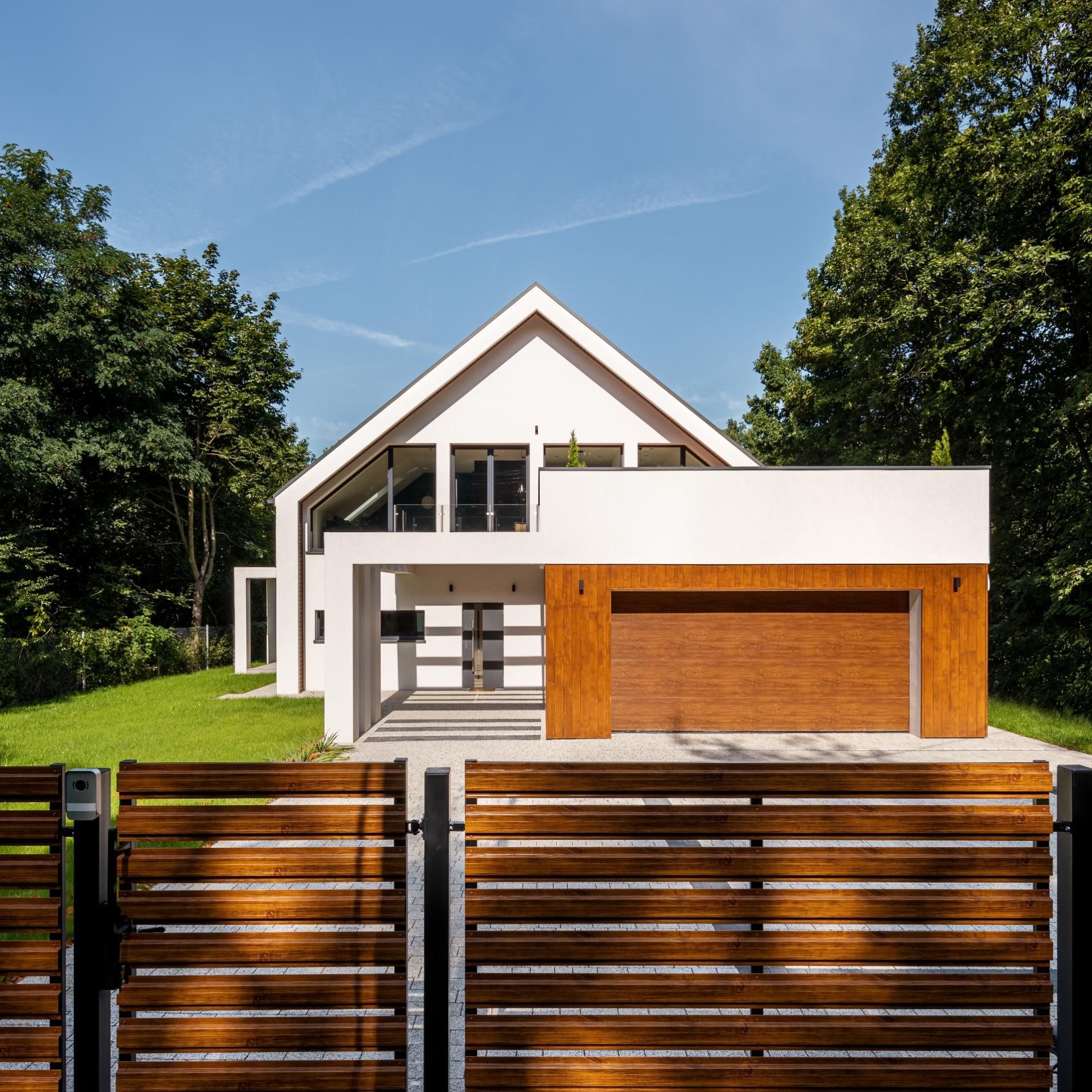 Modern white house with a wood garage door, wooden fence, and trees against a blue sky.