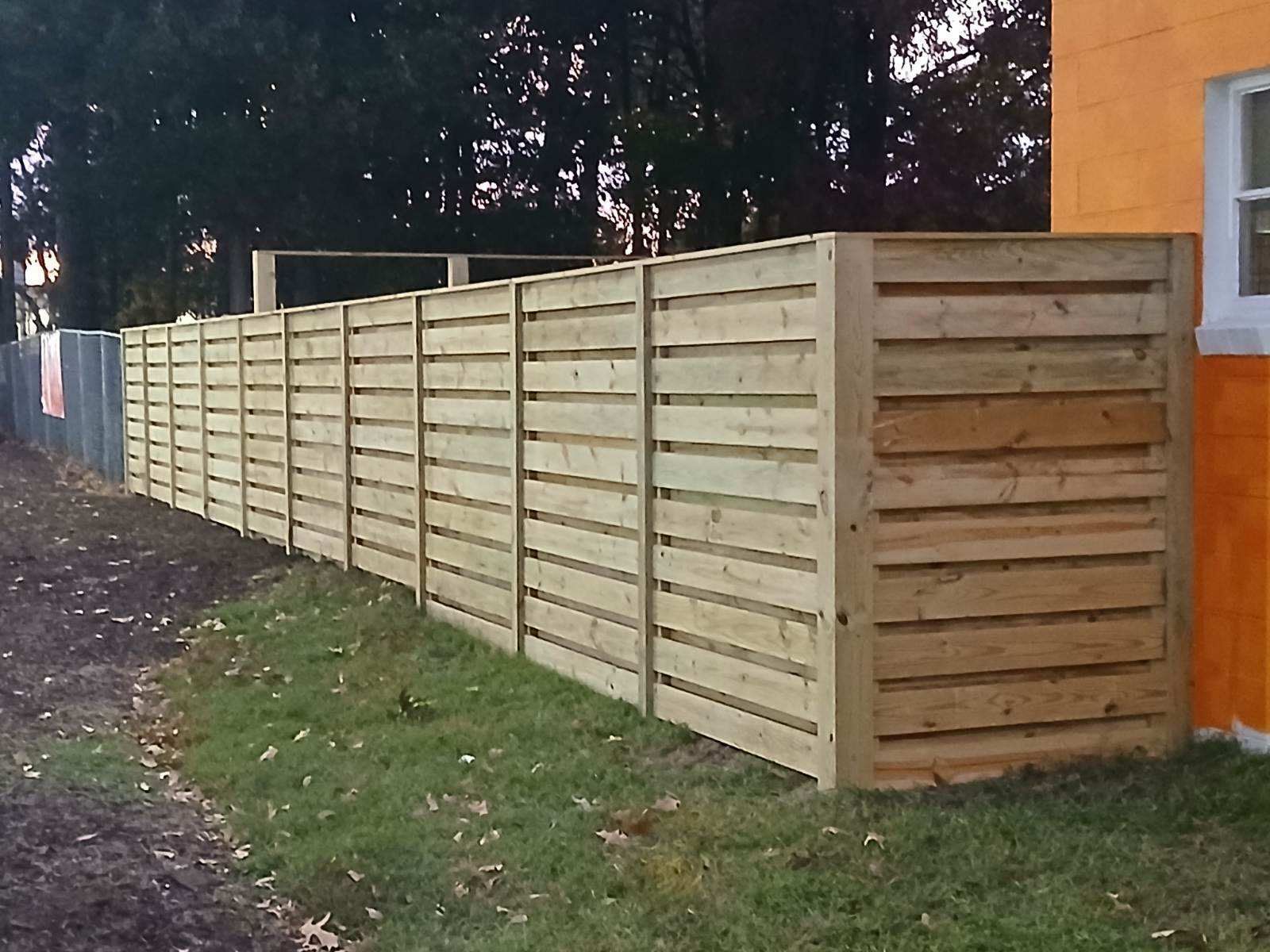 White picket fence bordering a yard next to a sidewalk and road, with green shrubbery in the background.