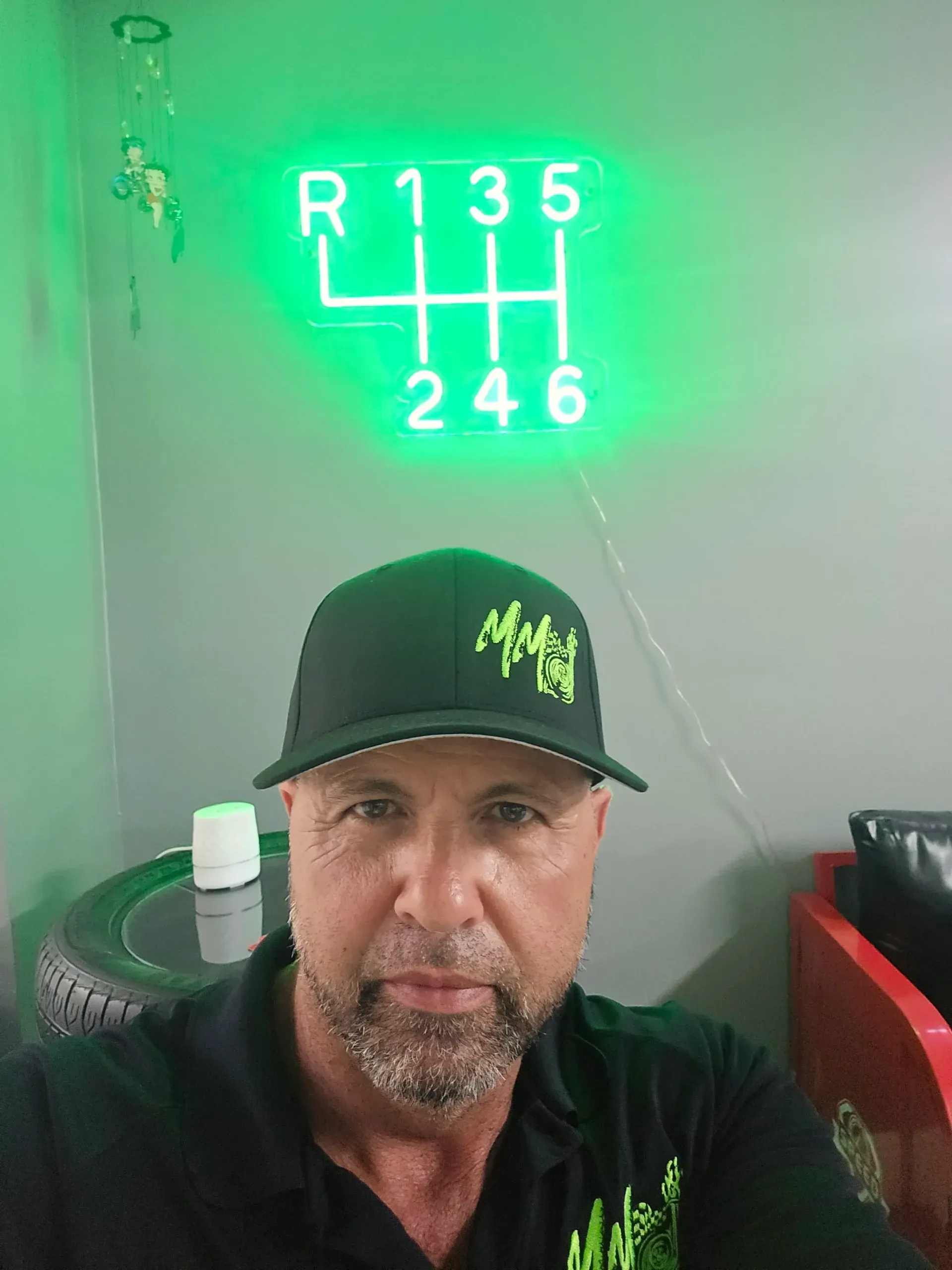 A person wearing a black baseball cap with lime-green logo sits before a neon wall sign showing a six-speed gear pattern.