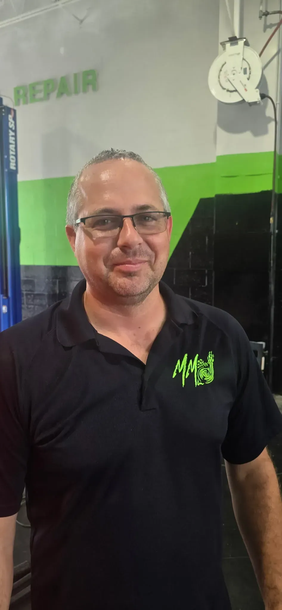 Smiling man in black polo standing in front of a green and black repair shop banner