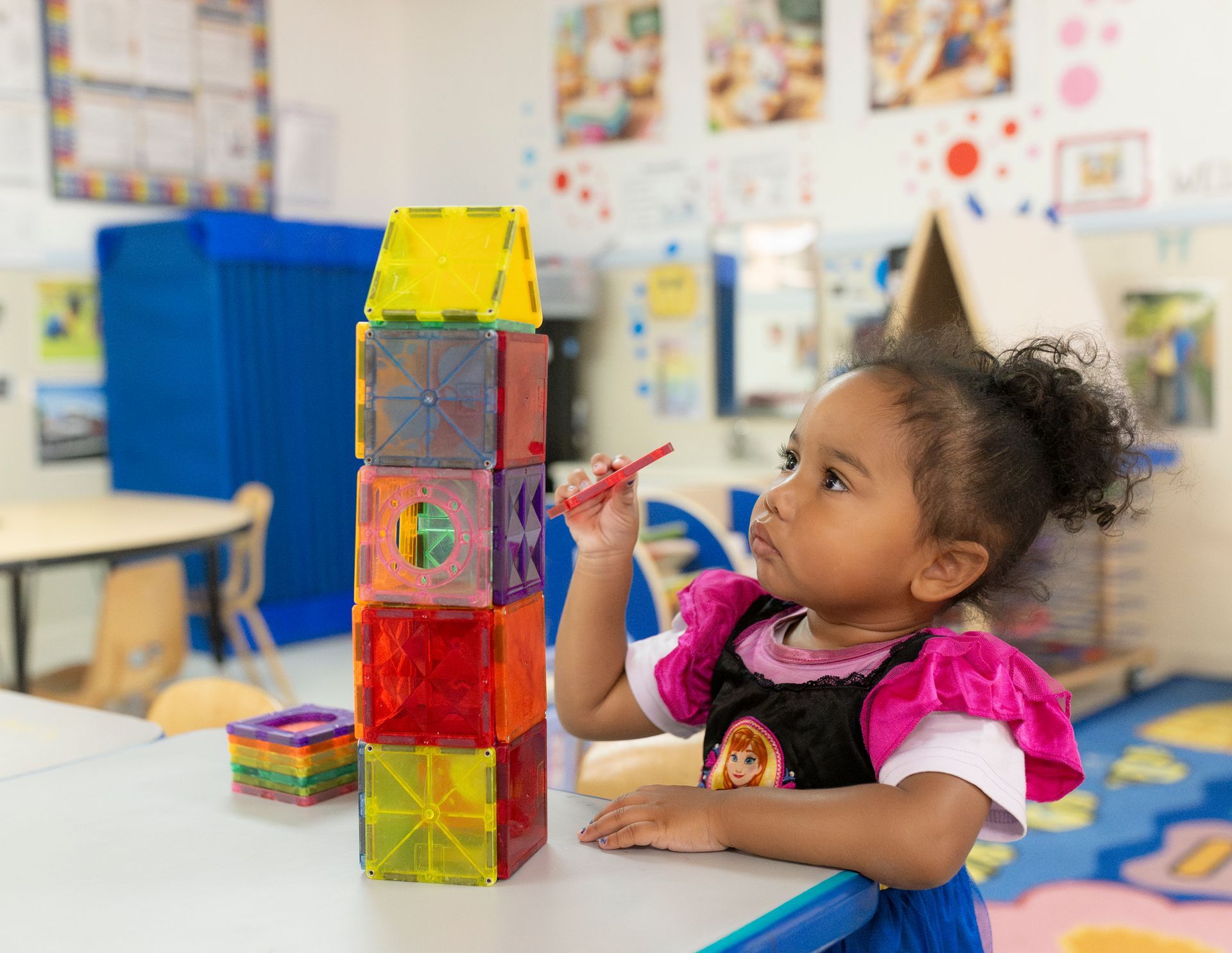 Child building a colorful block tower in a classroom, reaching with a red crayon.
