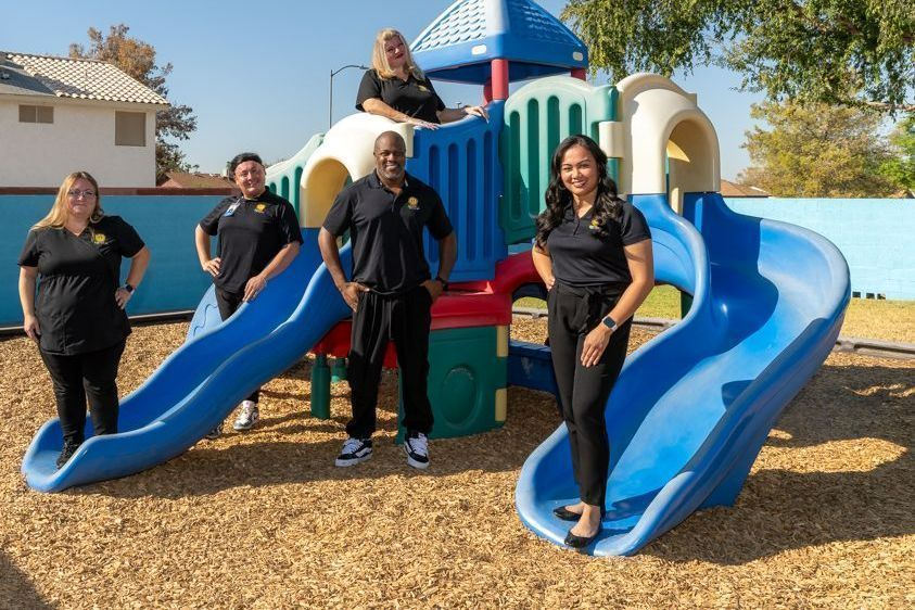 Five people posing on a playground slide structure with blue slides and wood-chip ground in a sunny park