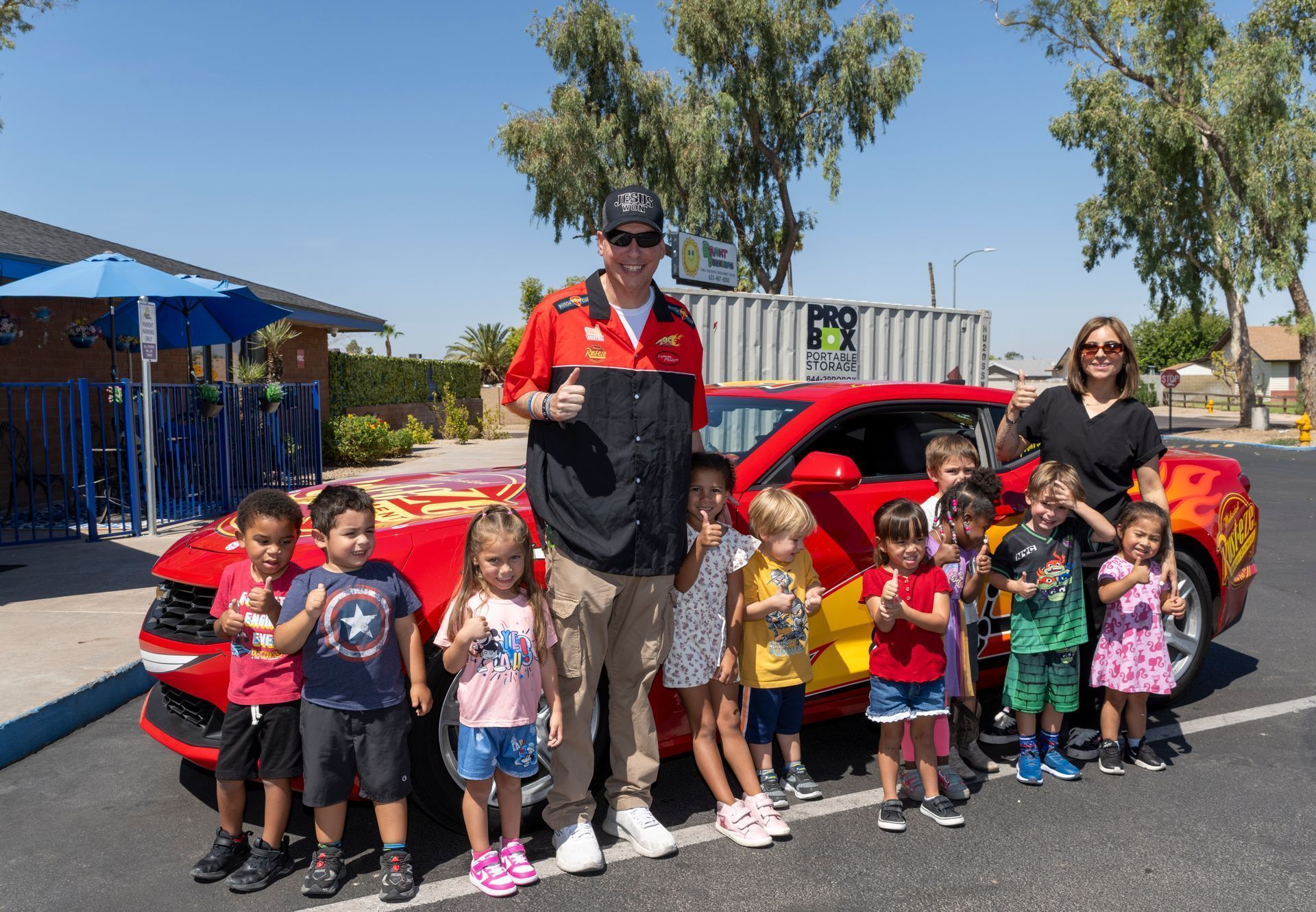 Group standing beside a red race car with a driver, outdoors on a sunny day.
