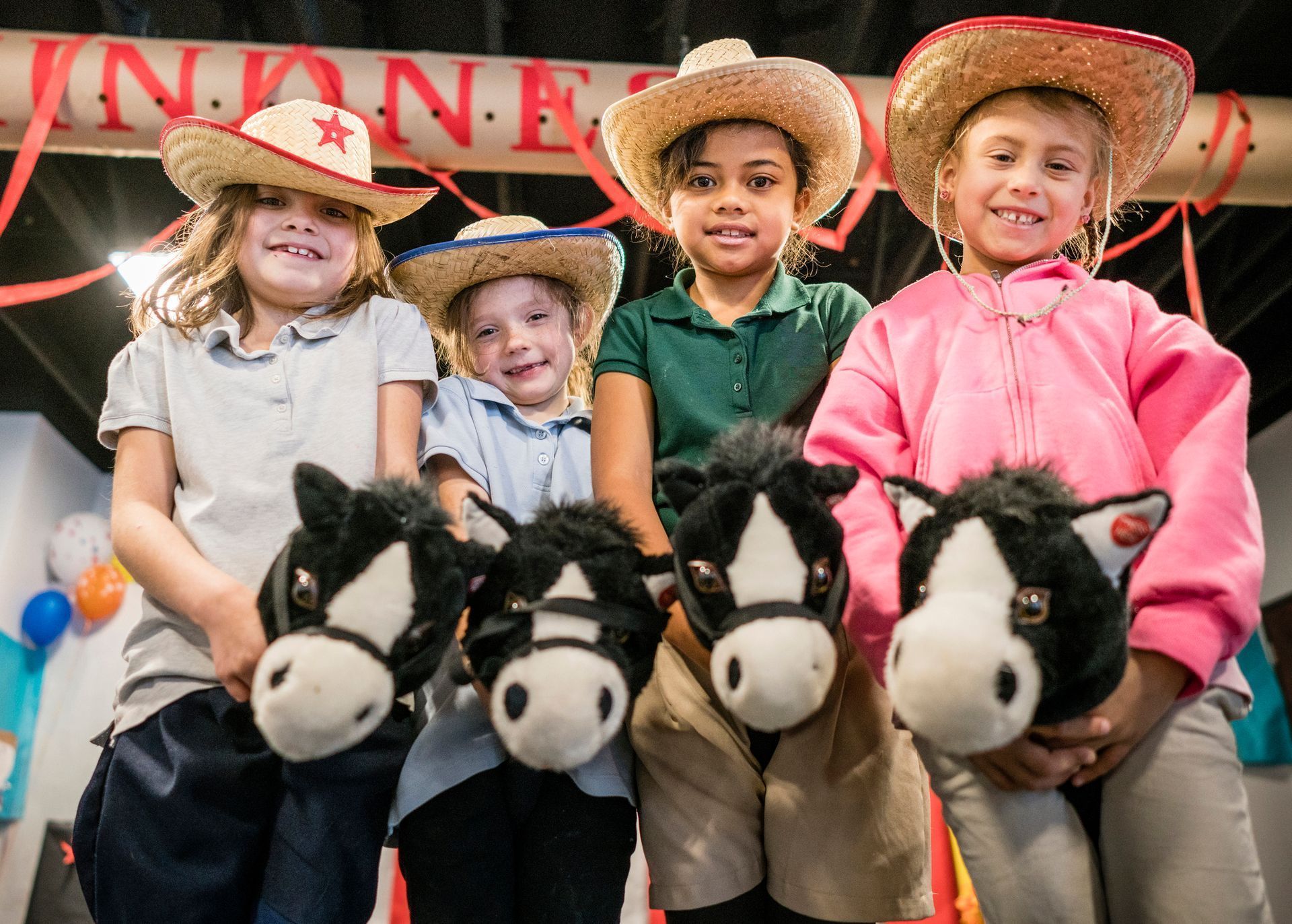 Four children in cowboy hats sit on toy horses, smiling at a rodeo-style event.