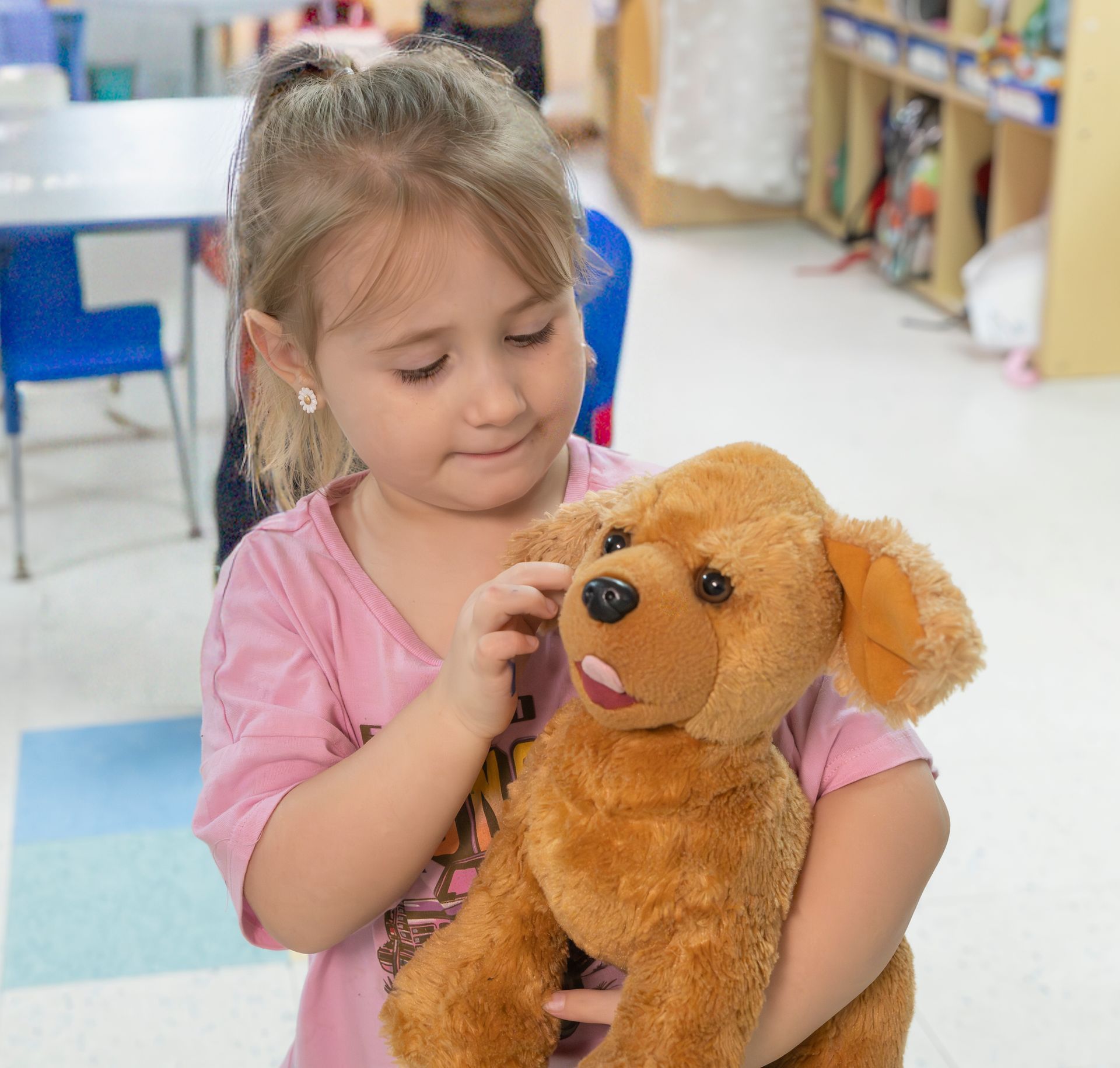 Young child in a pink shirt gently holding a brown stuffed dog in a classroom