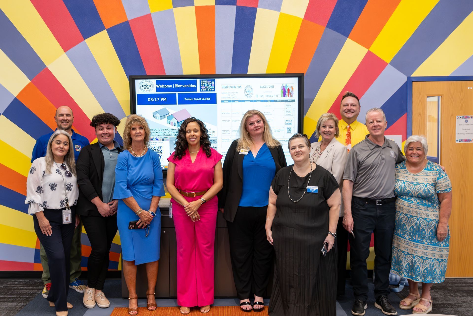 Group of people standing in front of a colorful mural and display screen at an indoor event