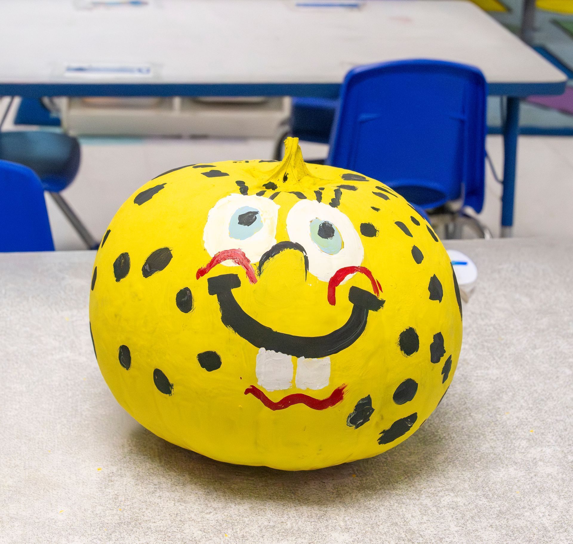 Yellow painted pumpkin with a smiling face on a classroom table, with blue chairs in the background.