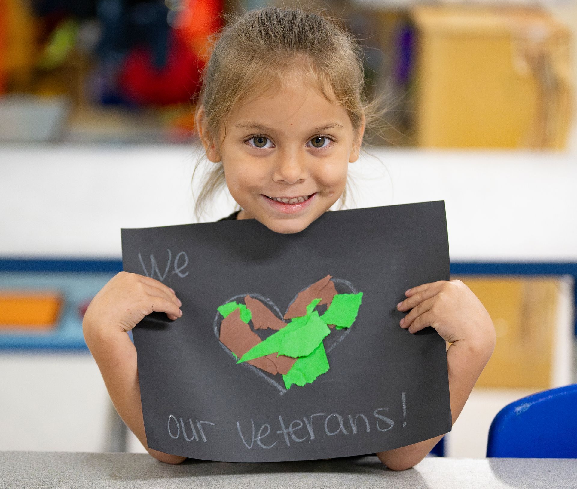 Child holding a black sign with a colorful heart and “We our veterans!” in a classroom setting.