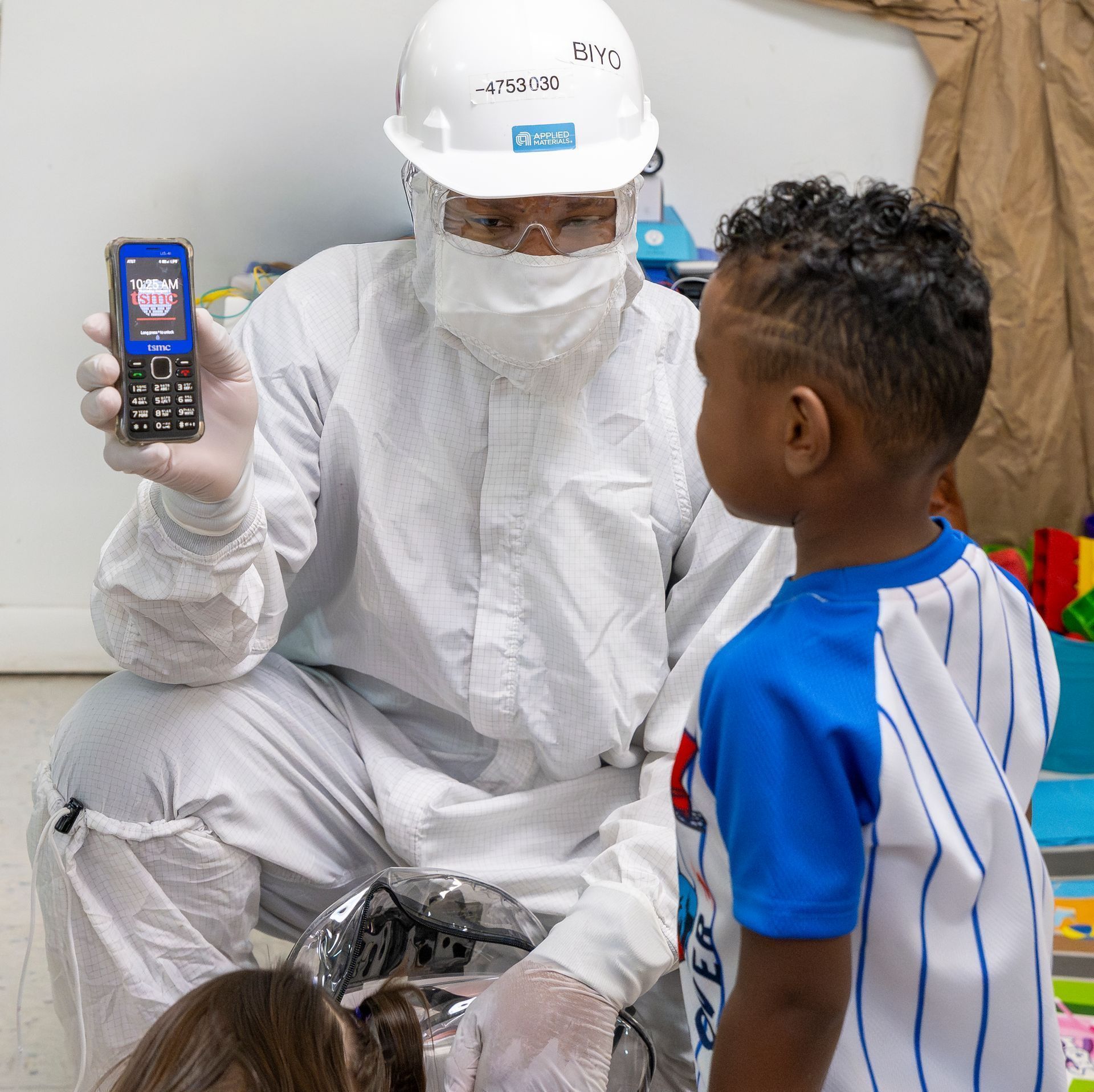 Health worker in white protective gear shows a phone to a child in a medical setting.