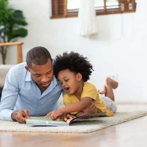 Adult and child lying on a rug, reading a colorful book together in a bright room