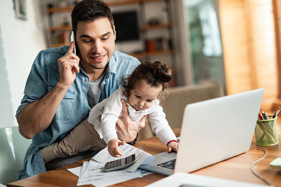 Man on phone helping a toddler use a calculator beside a laptop at a home desk