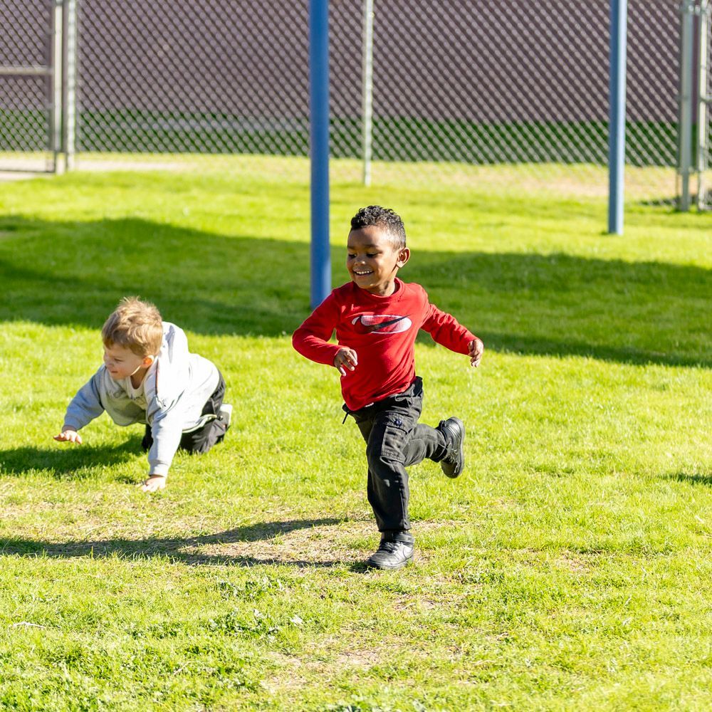 Two children running on a sunny grassy field near a fence