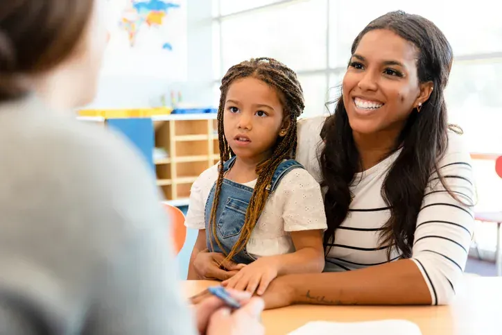 Woman smiling beside child in a classroom, listening to someone across the table