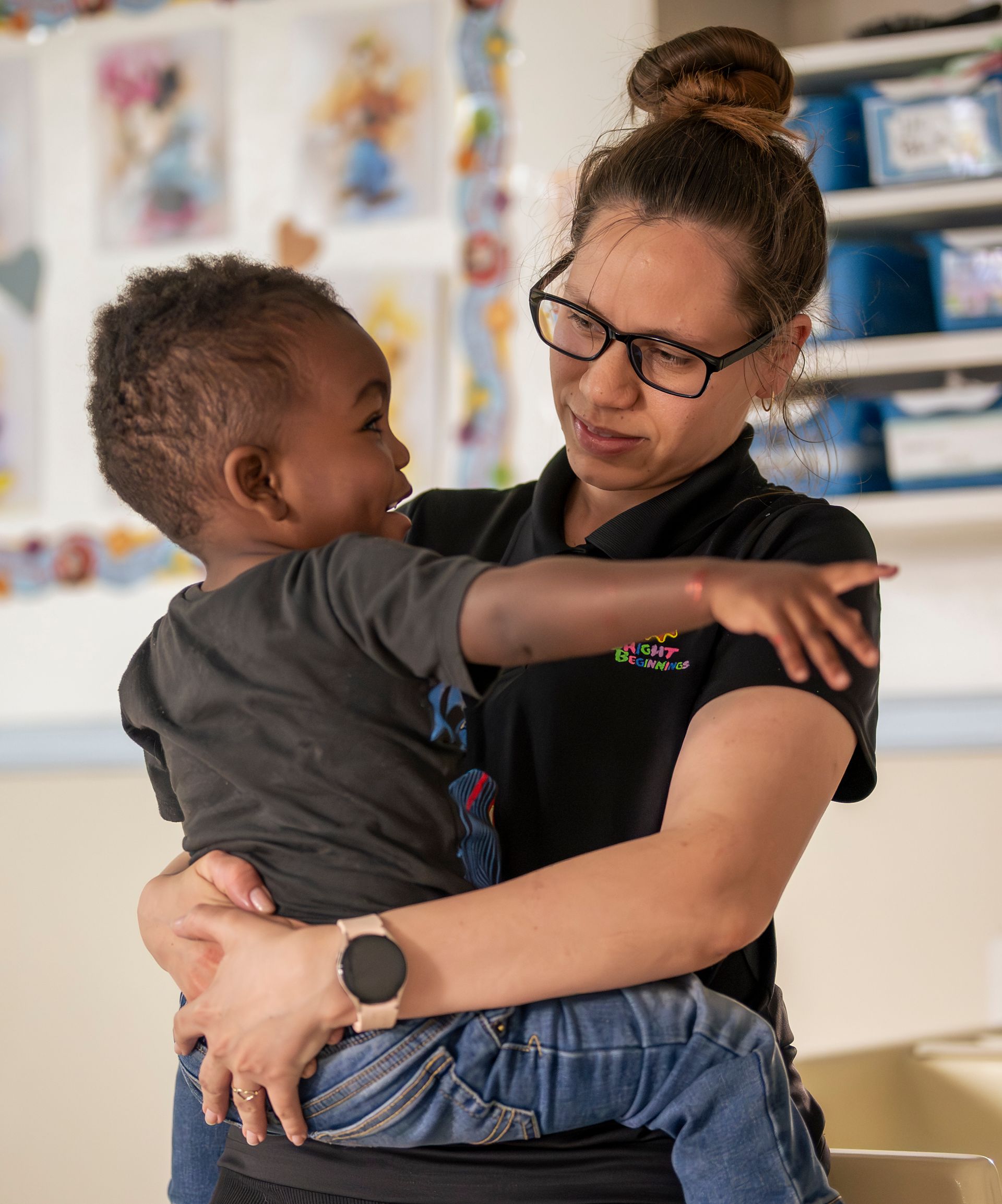 Woman holding a child in a classroom, both looking at each other with arms around one another