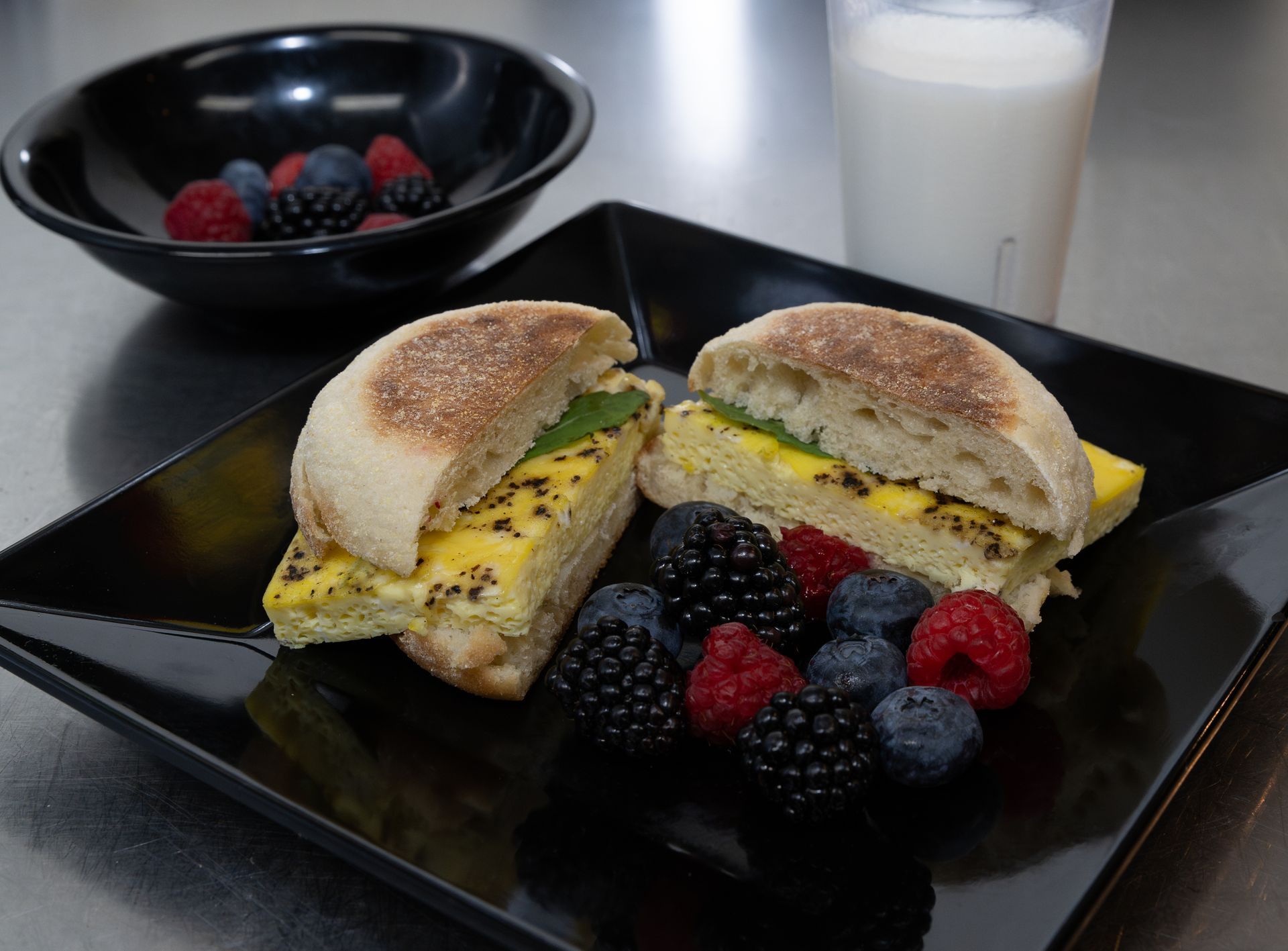 Breakfast sandwich on a black plate with berries and a glass of milk