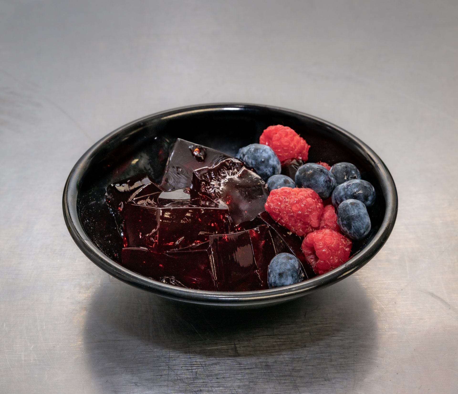 Black bowl of red jelly topped with blueberries and raspberries on a gray surface