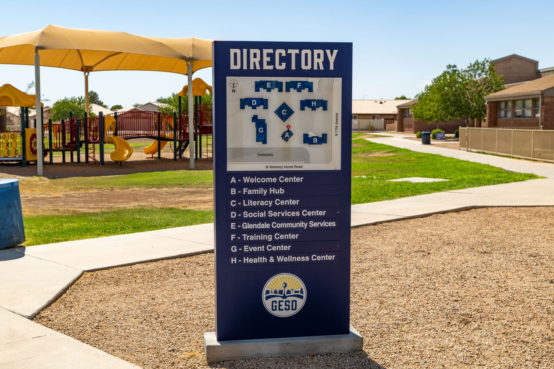 Outdoor directory sign at a sunny park with playground and apartment buildings in the background