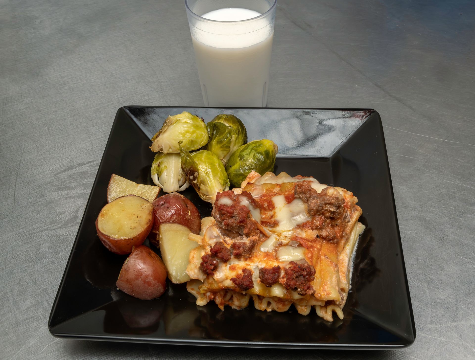 Plate of lasagna with roasted potatoes and Brussels sprouts, plus a glass of milk on a gray table