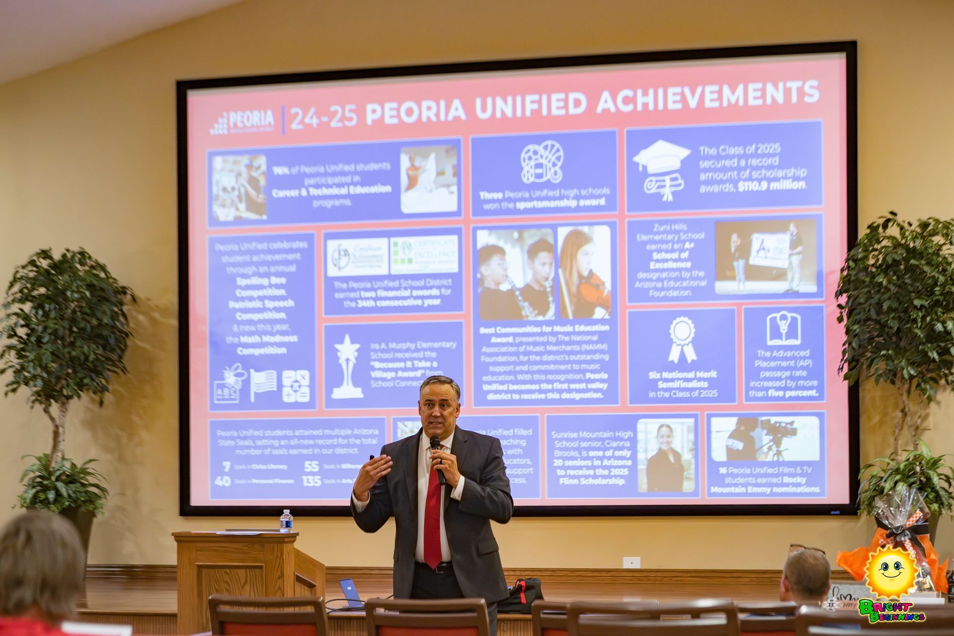 Speaker in a suit presents beside a large “Rejioa Unified Achievements” slide in a lecture hall