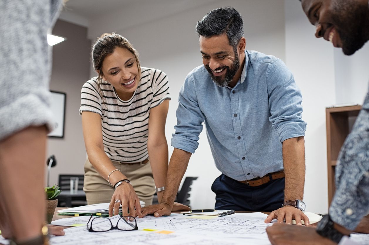 Three coworkers lean over a table, smiling and reviewing plans in a bright office.