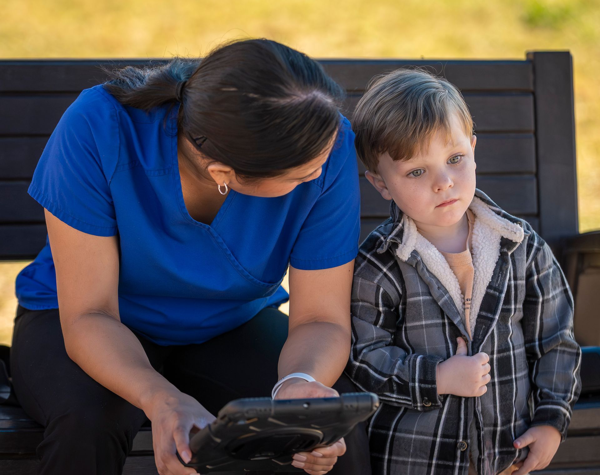 Woman and child sitting on a bench, looking at a tablet together outdoors.