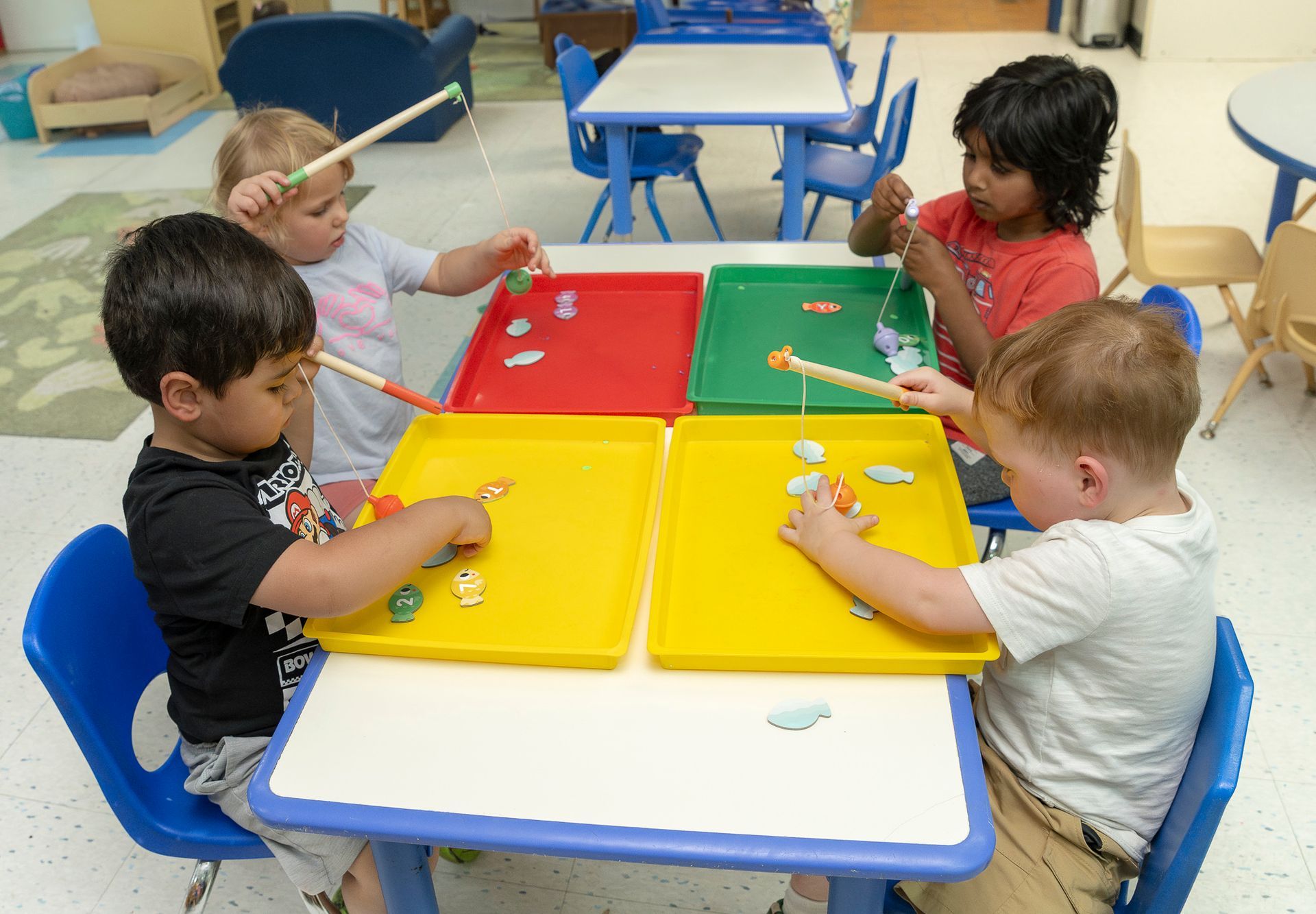 Four children painting at a low table with colorful trays in a classroom