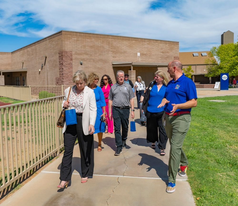 People walking on a sunny sidewalk beside a brick building, with one man in a blue shirt speaking to the group.