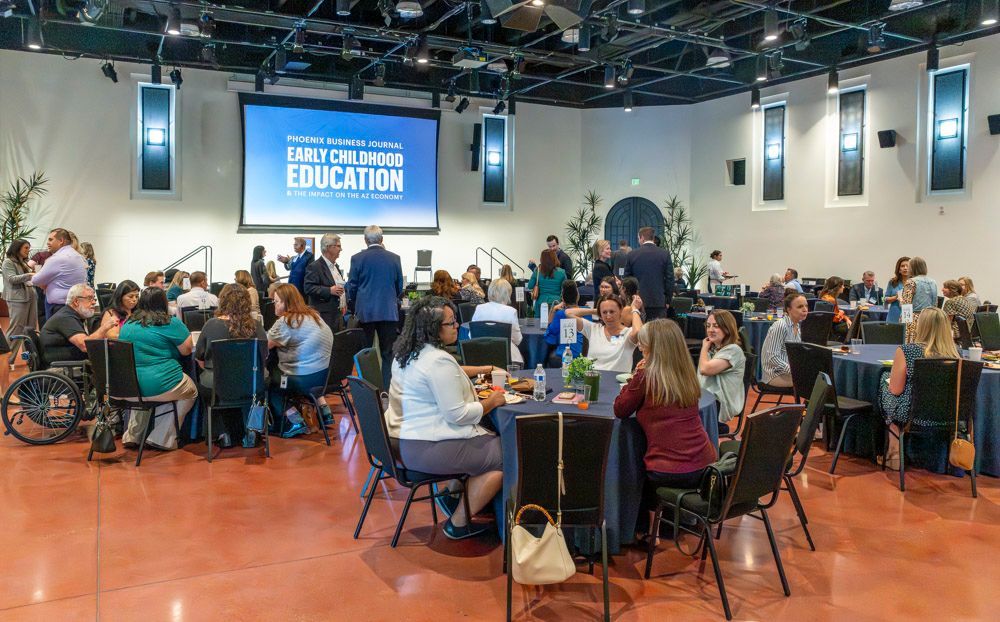 Conference attendees seated at round tables in a large hall, facing a screen that reads “Community Education.”