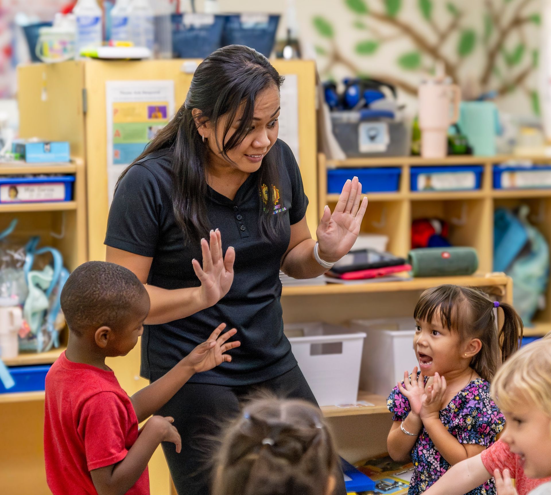 Teacher leading a classroom activity with children, hands raised and smiling, in a colorful preschool room