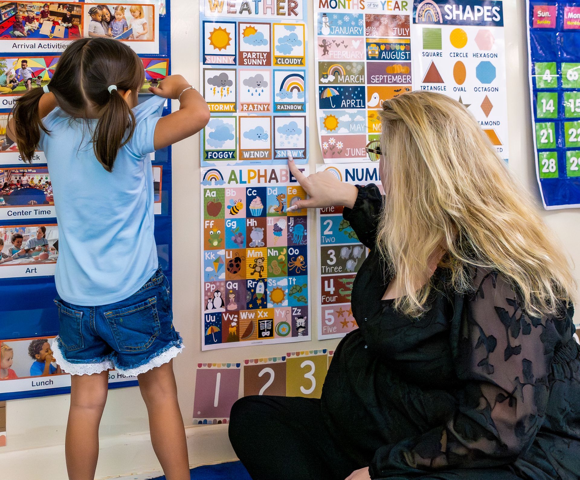 Teacher and child pointing at colorful classroom learning charts on a wall