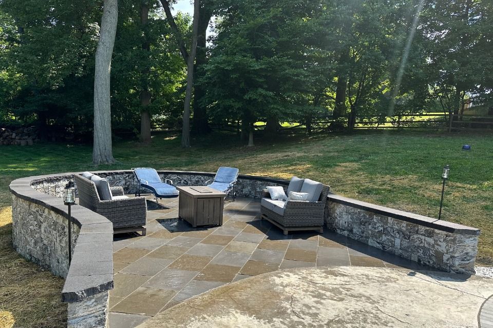 A patio with a couch , chairs , and a fire pit surrounded by trees.
