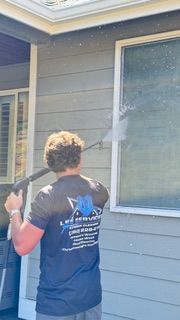 Person in yellow rain gear power washing a gray house with white-framed windows, outdoors.