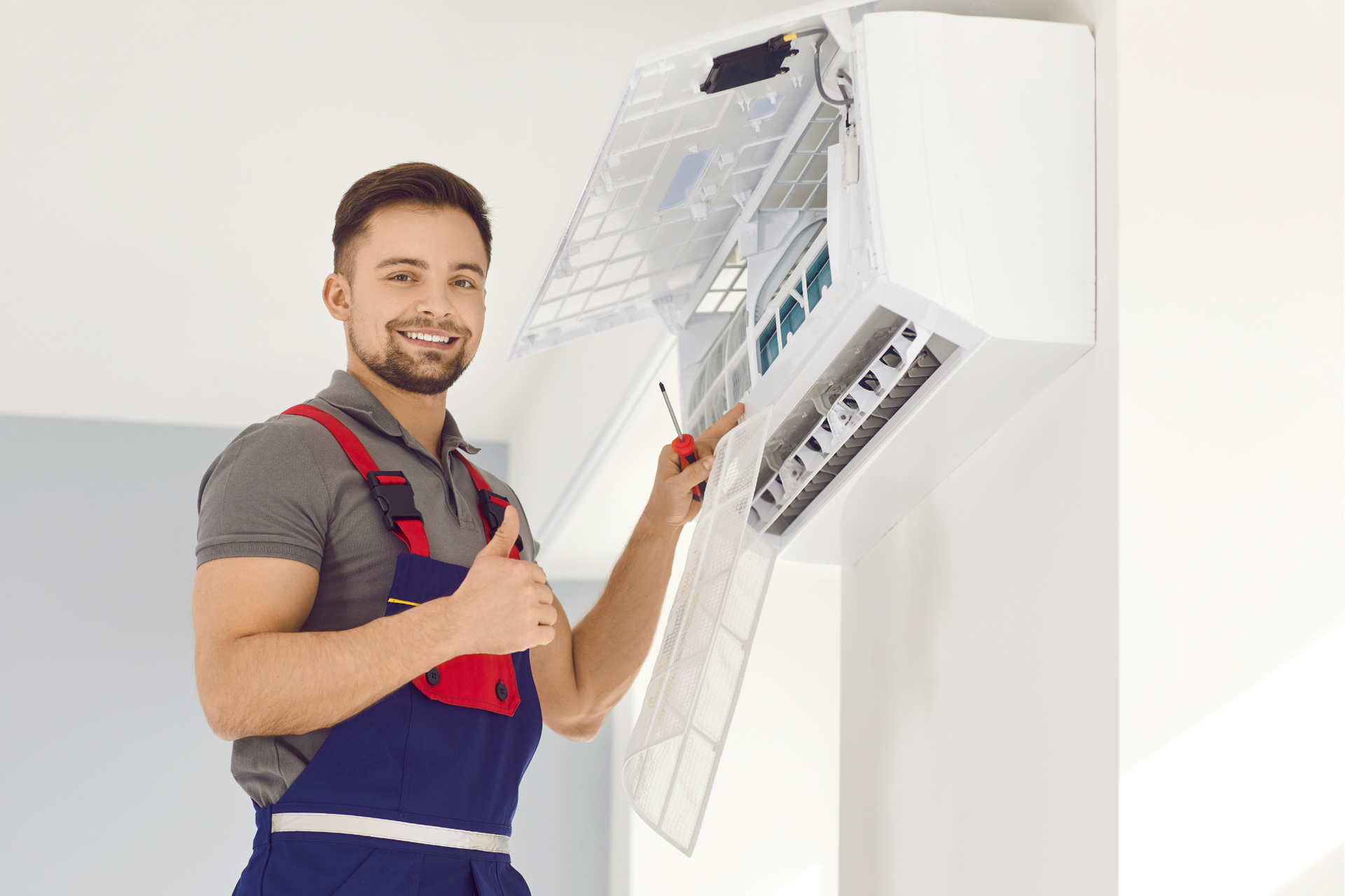 A man is fixing an air conditioner with a screwdriver and giving a thumbs up.
