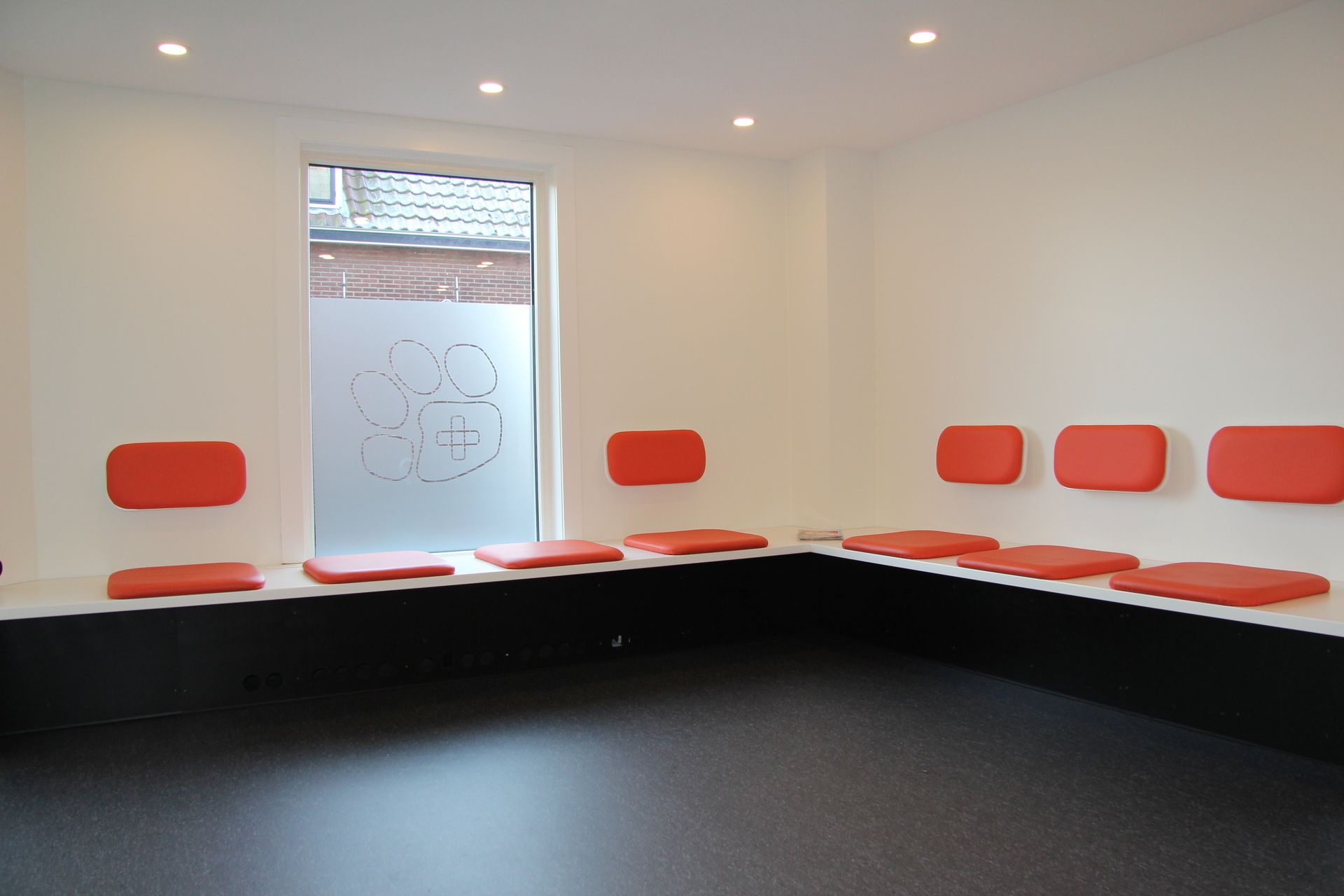 Lege wachtkamer met rode stoelen langs witte muren en een raam, boven een zwarte vloer.