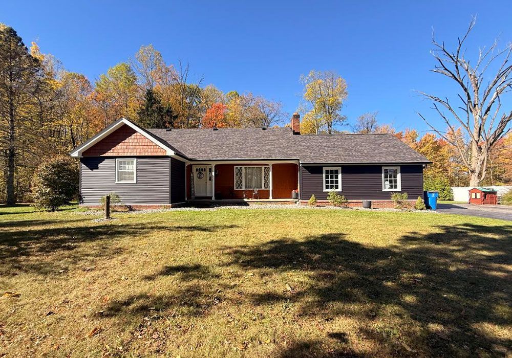 Dark blue house with brown roof and red door on a grassy lot, surrounded by trees.