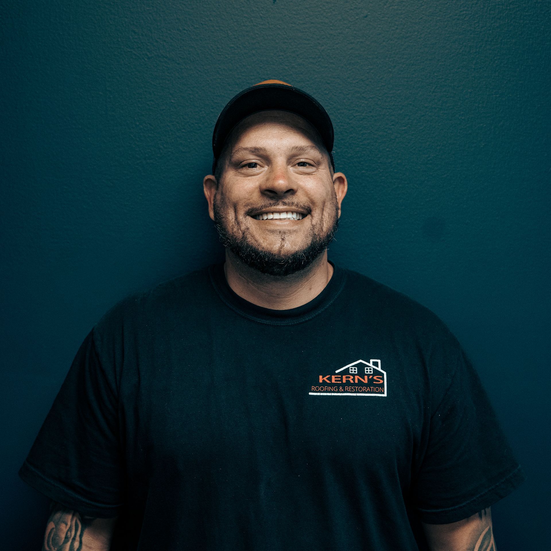 Smiling man in a black cap and T-shirt standing against a teal wall